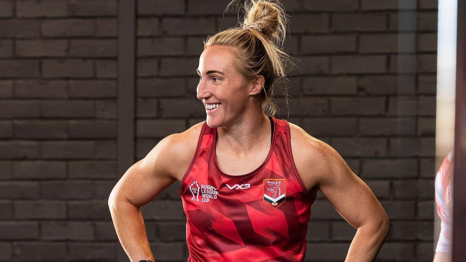 Bethan Dainton in red vest smiles in the gym in front of a brick wall